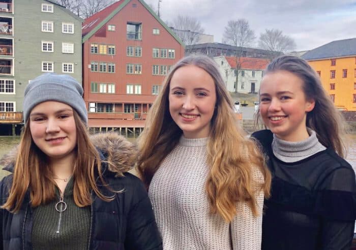 Drei lächelnde Austauschschüler:innen vor bunten Holzhäusern am Fluss in Skandinavien