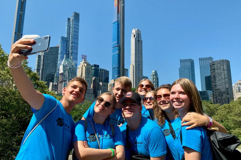 Austauschschüler:innen in blauen T-Shirts machen ein Selfie vor Wolkenkratzern in New York