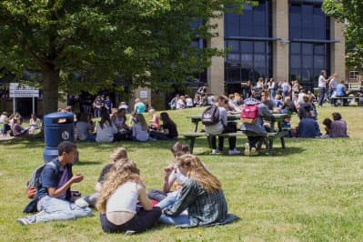 Schüler:innen sitzen bei Sonnenschein auf dem Rasen und an Picknicktischen vor ihrer Schule in Südostengland