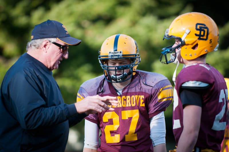 Footballtrainer spricht mit zwei Spielern in Helmen und Trikots – Teamsport im Auslandsjahr