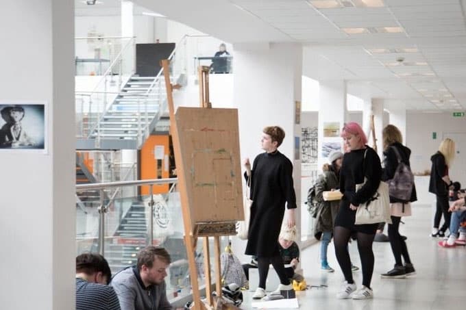 Schülerinnen im hellen Atrium neben einer Staffelei mit Zeichnung – Schulalltag in England