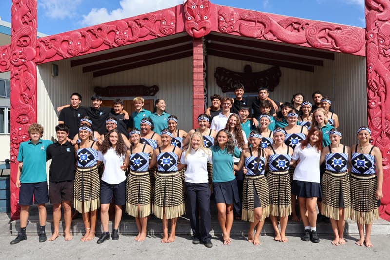 Schüler:innen in Māori-Tanzkostümen vor einem geschnitzten Versammlungshaus in Neuseeland